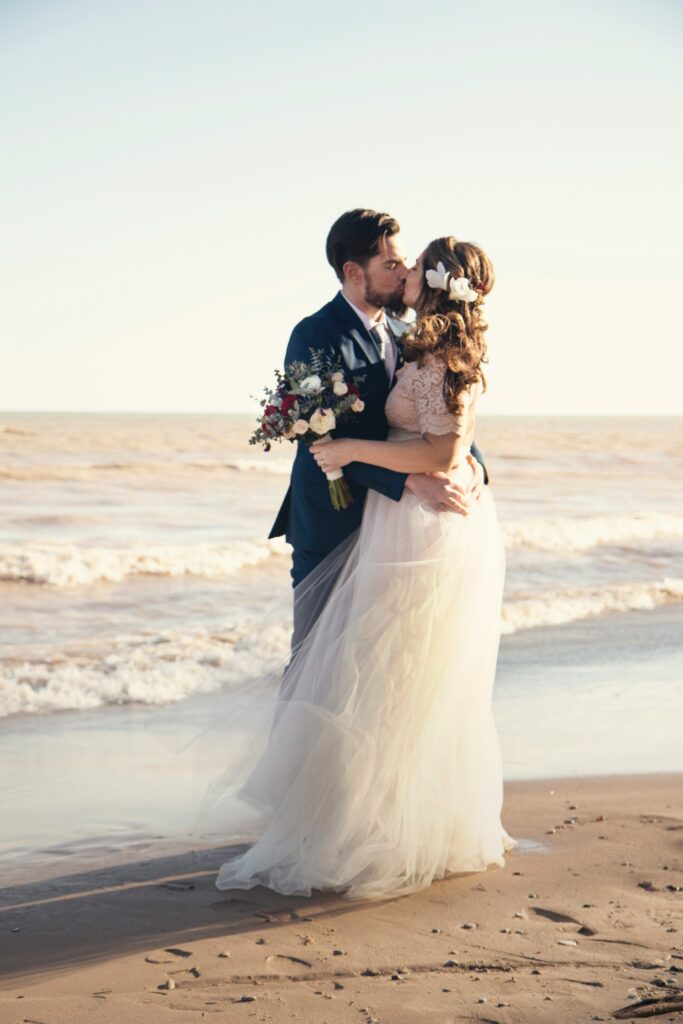 "Wedding couple in elegant bridal gown and tuxedo kissing on a serene beach. Explore bridal gowns and tuxedos for magical moments"